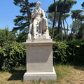 Estatua de mármol de Lord Byron sentado sobre un pedestal, en un jardín rodeado de árboles y vegetación. En el pedestal aparece la inscripción «BYRON 1788–1824» junto a un fragmento de una de sus obras, Childe Harold